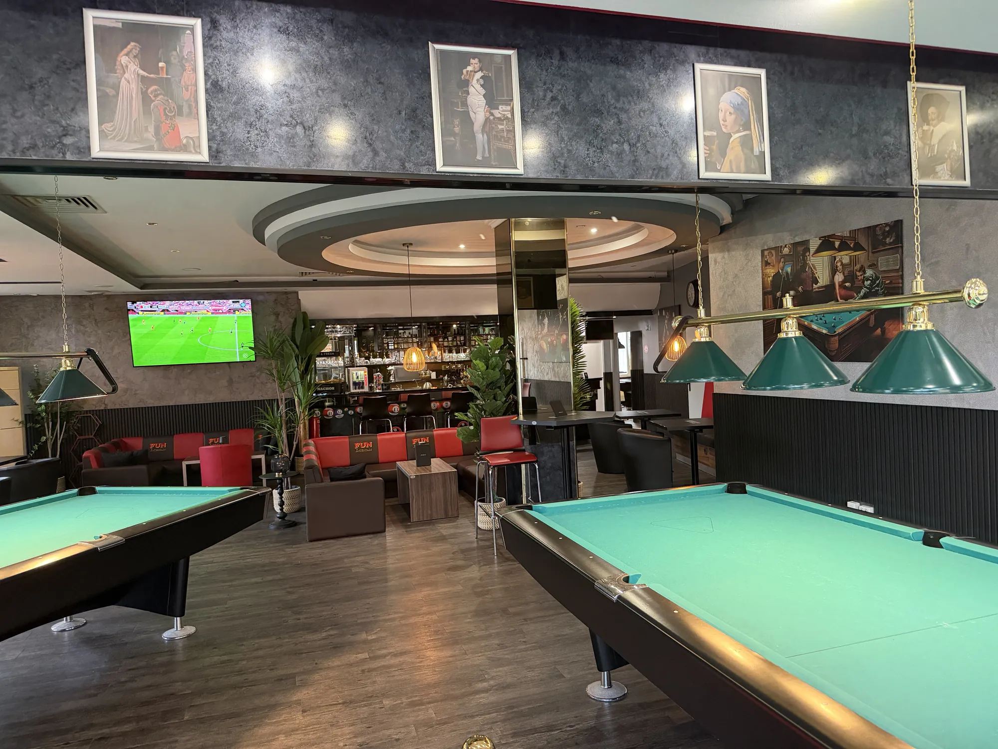 Zwei Poolbillardtische mit grünem Tuch unter Messinglampen in Dorsten, dahinter Sitzbereich mit roten Sofas und die Bar